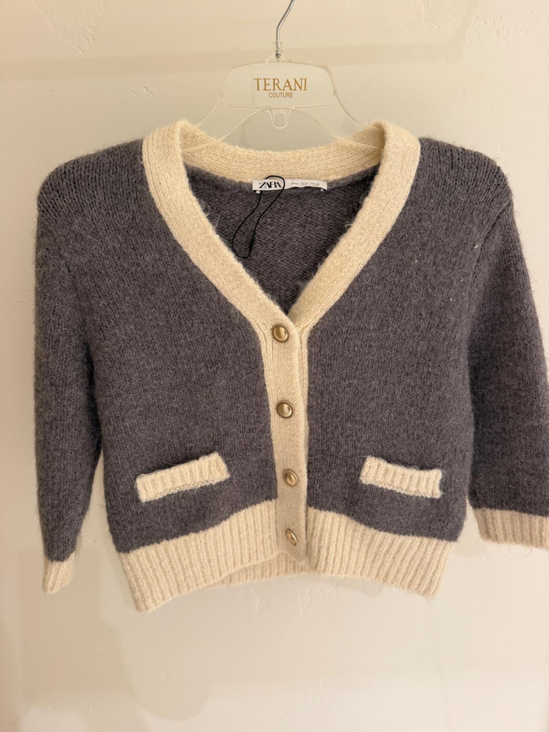 Grey cardigan with beige trim and gold buttons, hanging on a clear hanger with "TERANI COUTURE" text.