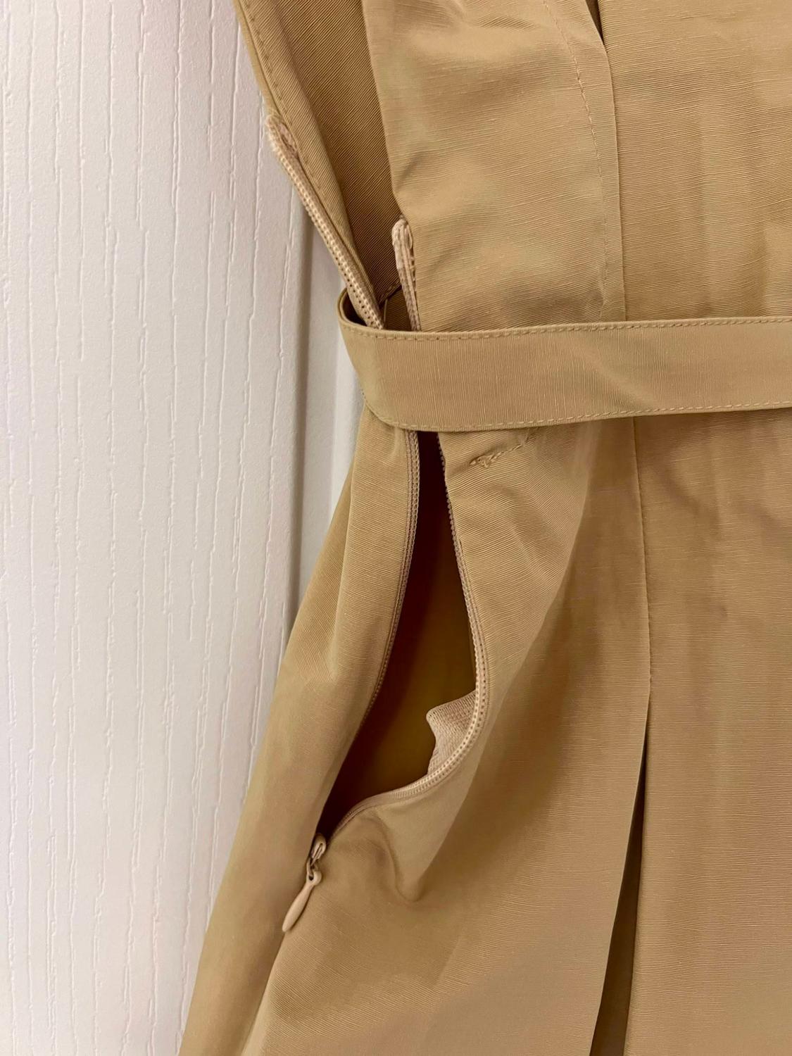 Detail of a beige garment with a matching belt and a semi-open side zip revealing a pocket, set against a white door background.
