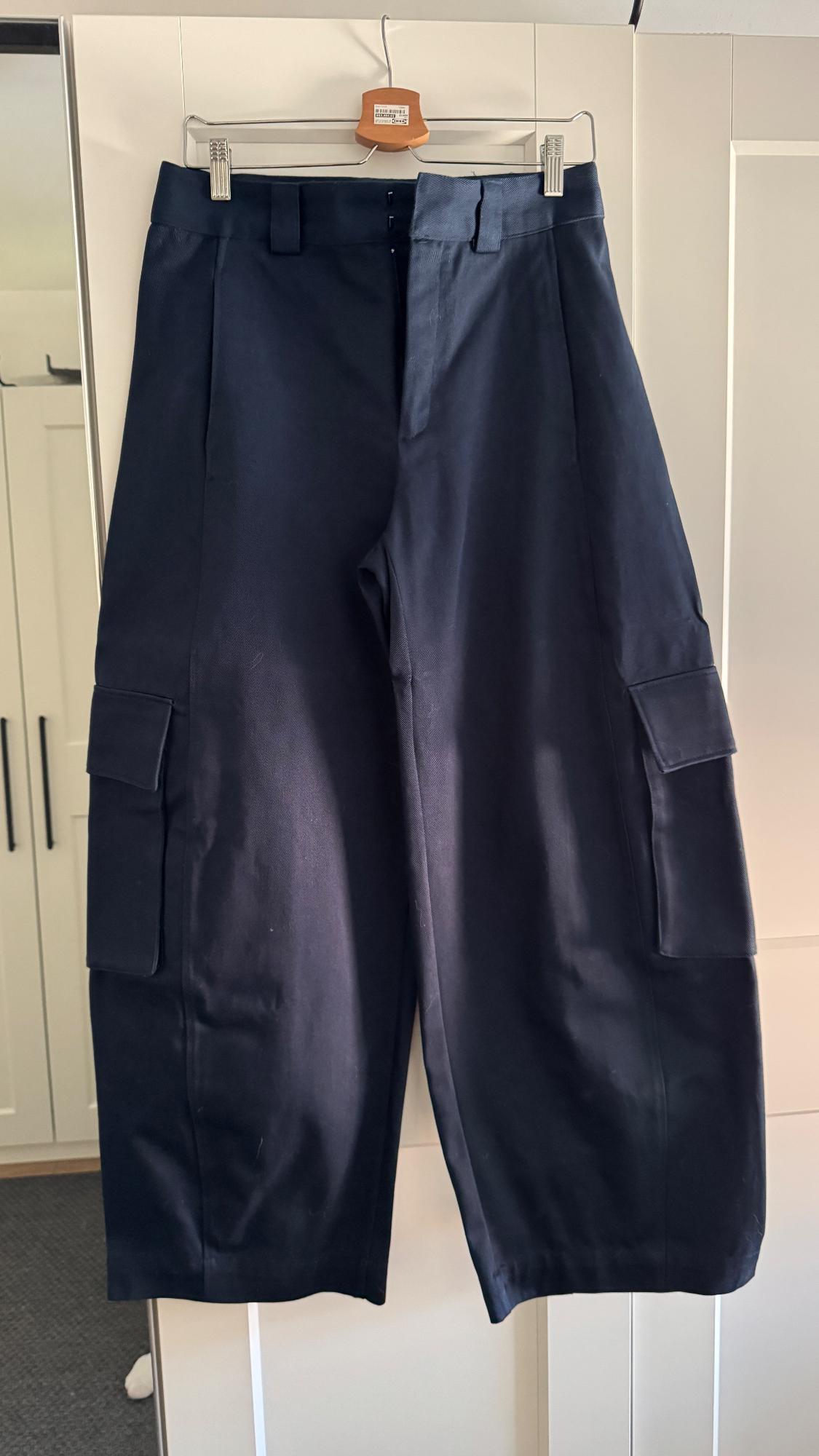 Navy blue trousers with large pockets hanging on a wooden hanger, white wardrobe background.