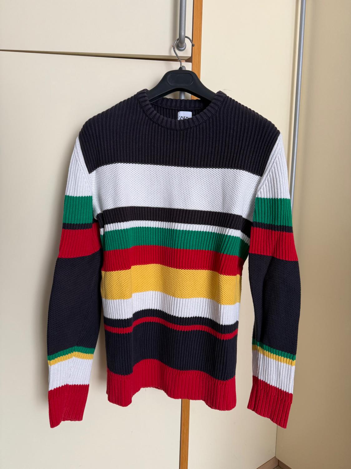 Multicoloured striped jumper hanging on a black hanger. Colours: black, white, red, green, and yellow. Beige wardrobe background.