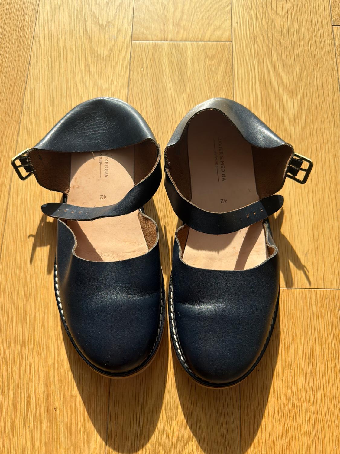 Black leather shoes with gold buckles on a light wood floor. Visible text: "JAVIER MEDINA" and "42".