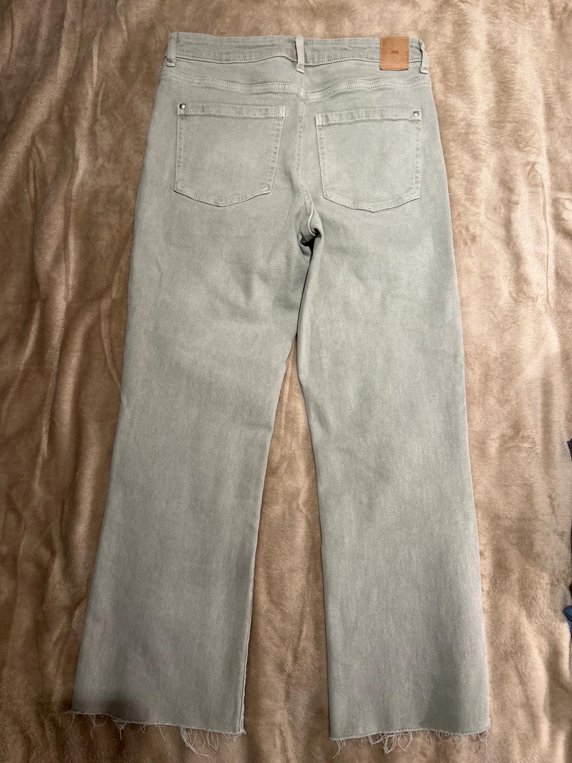 Grey denim jeans with a frayed hem, viewed from behind on a brown fabric surface. Brown label on the waistband.