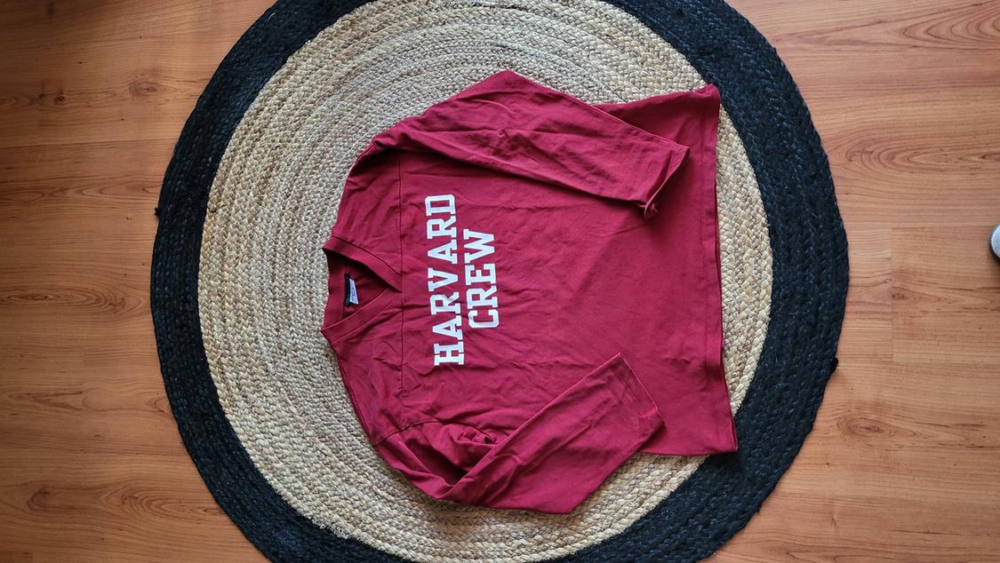 Red long-sleeved t-shirt with "HARVARD CREW" text in white, on a round jute rug on a wooden floor.