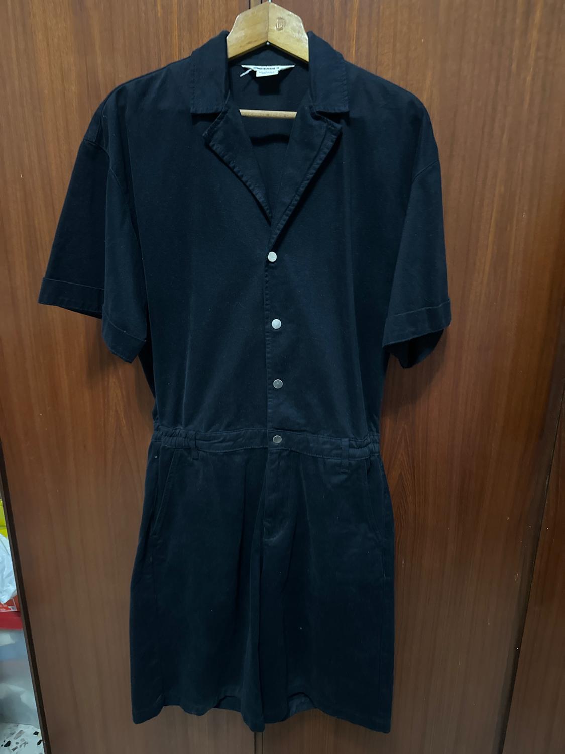 Black short-sleeved jumpsuit with silver buttons, hanging on a wooden hanger against a brown wooden background.