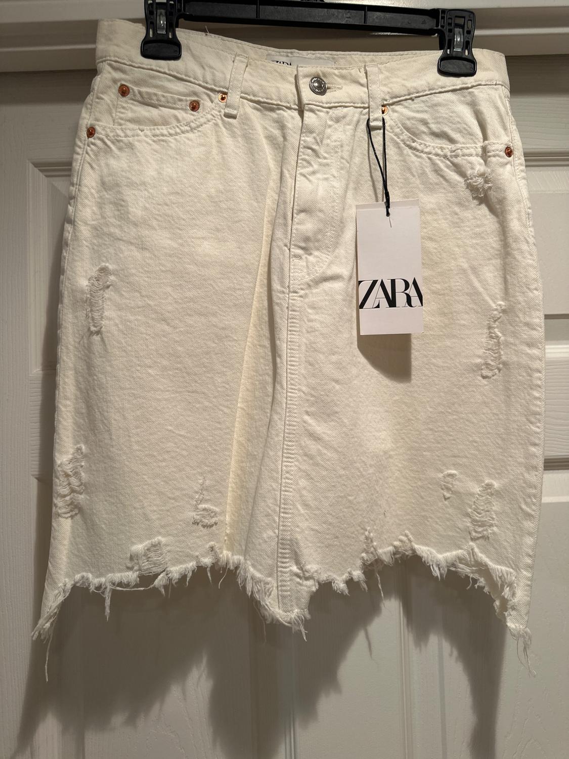 White denim skirt with frayed edges and distressed detailing, hanging on a hanger. Label with text "ZARA".