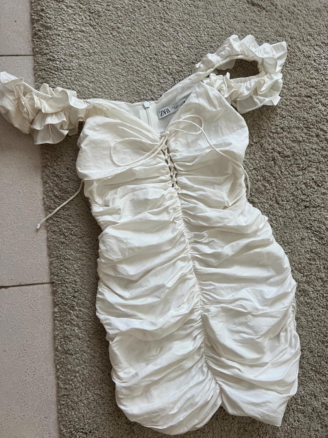 White ruched off-the-shoulder dress with frill sleeves and lace-up detail. ZARA label. Zenithal photograph on a beige rug.