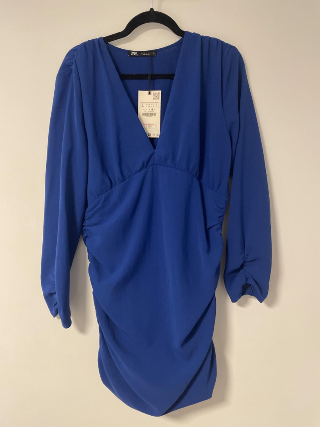 Electric blue dress with a V-neck and long sleeves, hanging on a black hanger. Label visible on the collar.