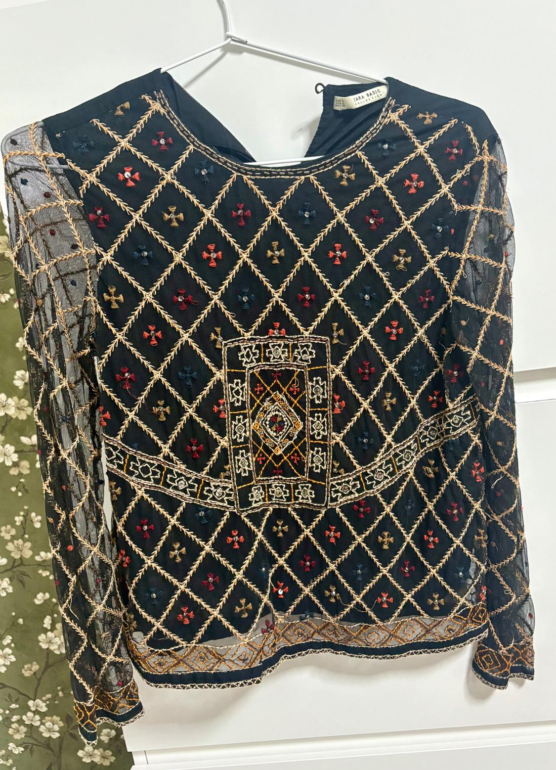 Black blouse with gold geometric embroidery and red and blue floral details, hanging on a hanger against a white background.