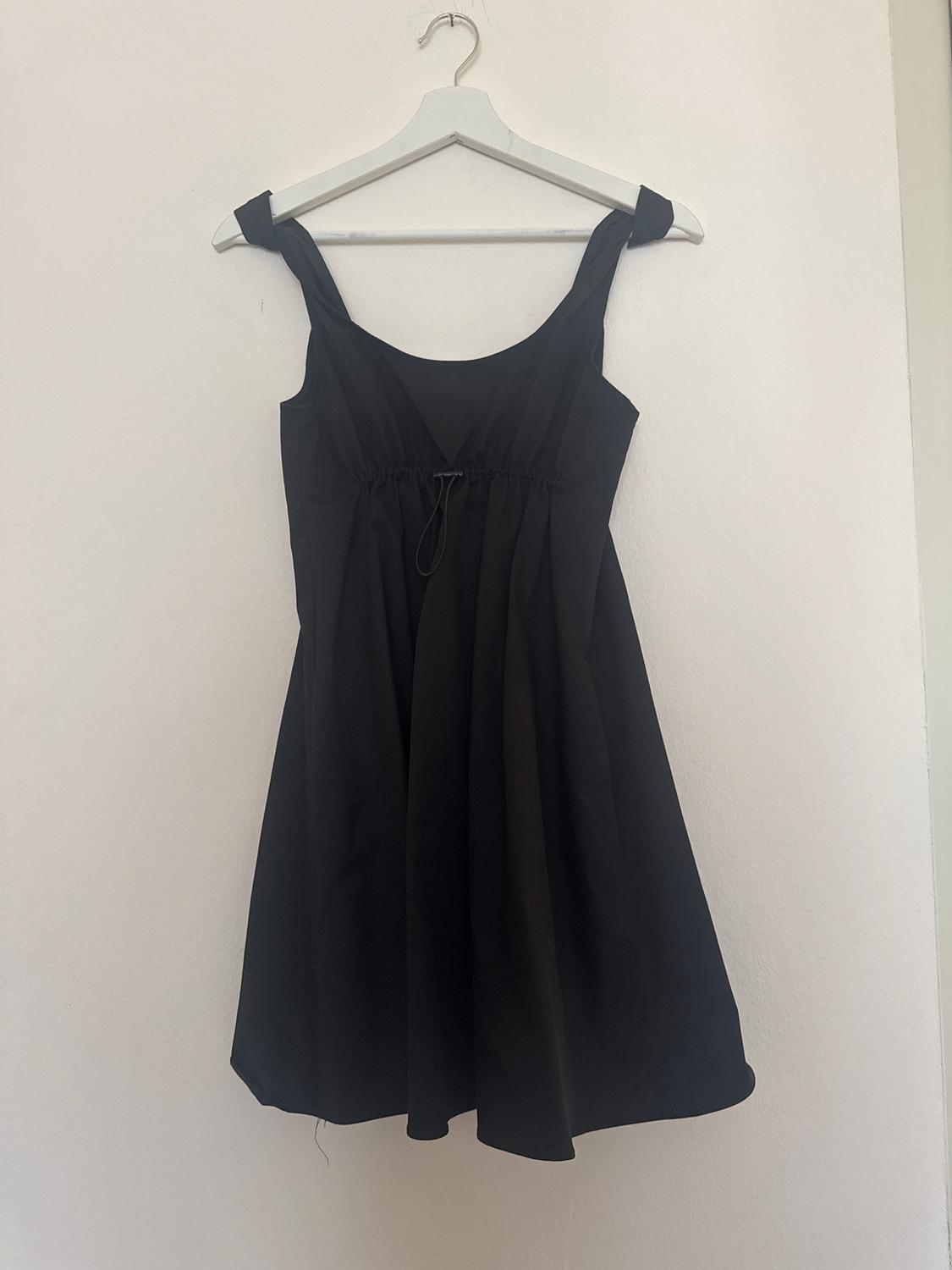 Sleeveless black dress hanging on a white hanger against a white wall. Simple design with wide straps.
