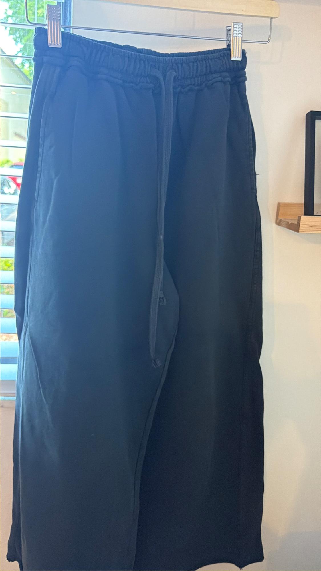 Dark pants with an elastic waistband and drawstring hanging on a hanger next to a window with blinds. Relaxed atmosphere.