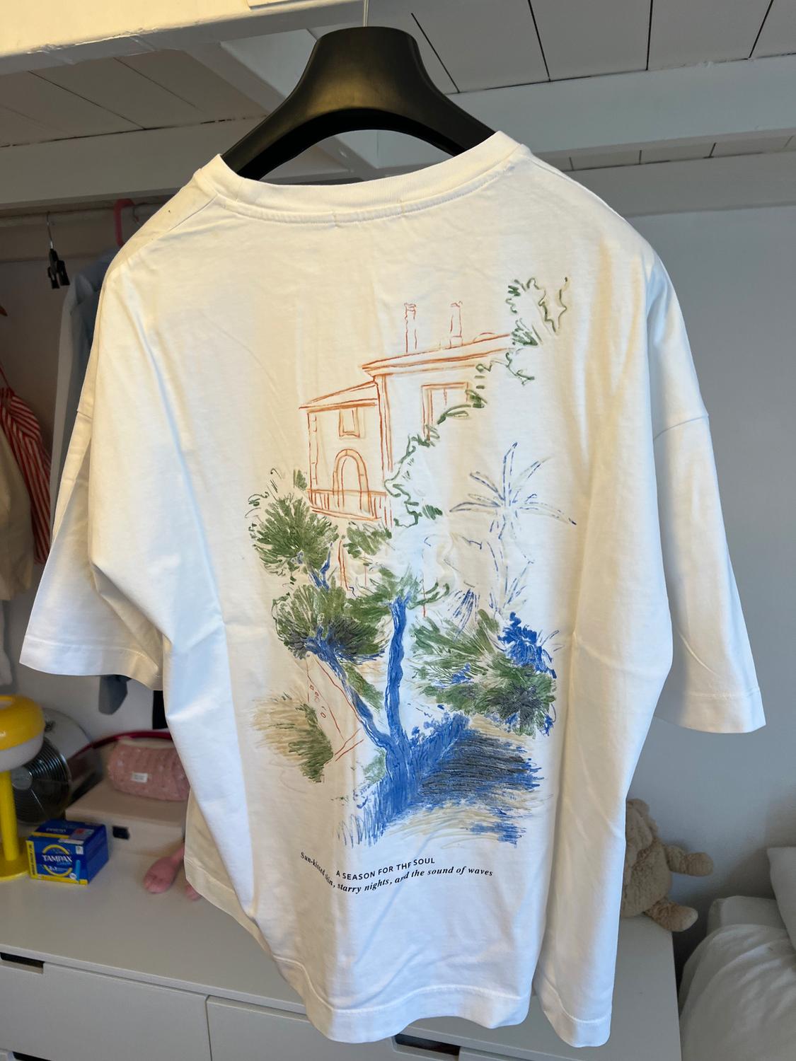 White t-shirt hanging with landscape illustration in green and blue colours. Text: "A season for the soul, starry nights, and the sound of waves".