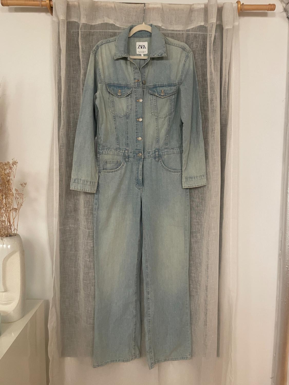 Light blue denim jumpsuit hanging on a hanger in front of a white curtain. Visible label with the text "ZARA".