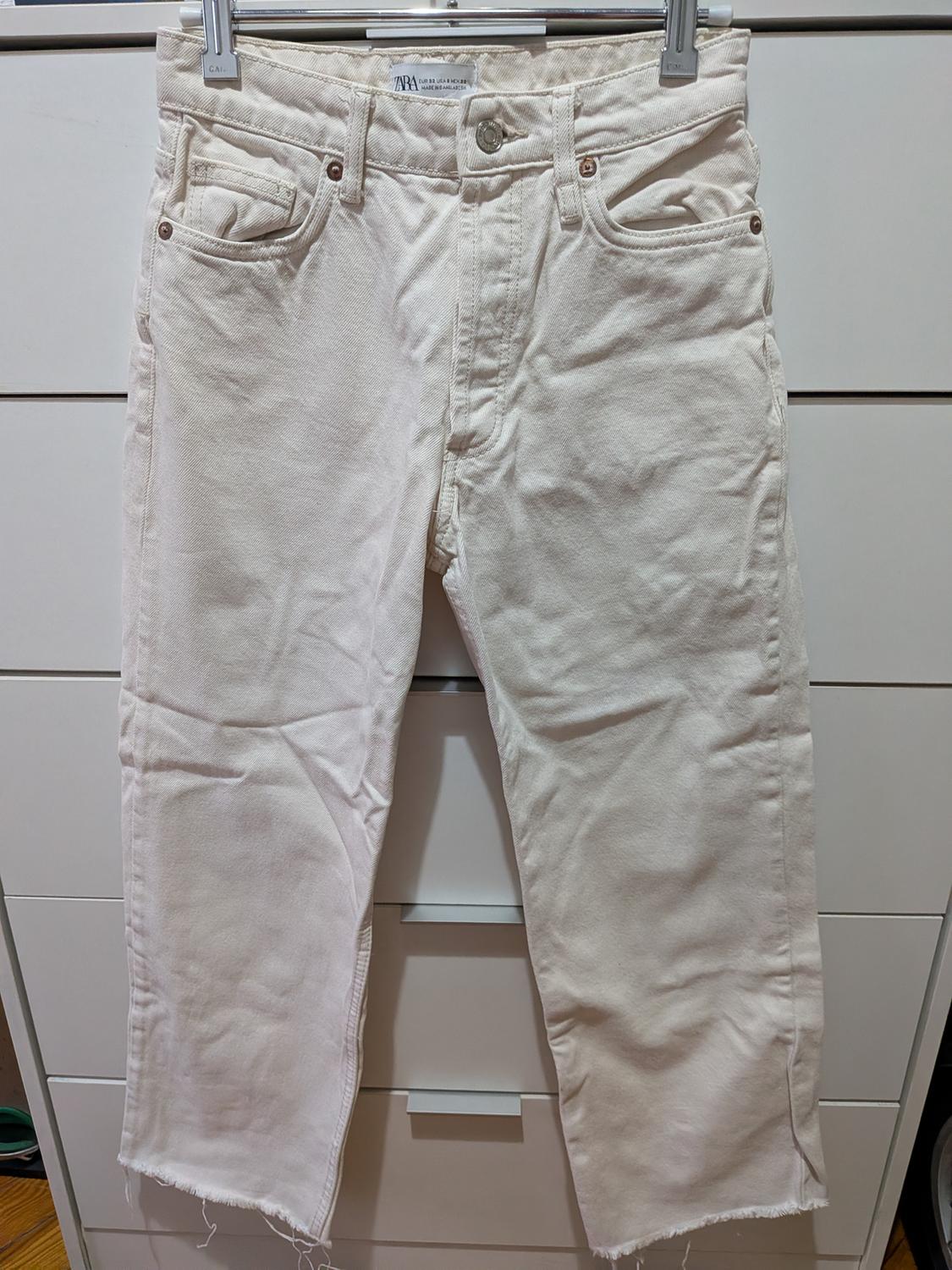 Beige denim jeans with a frayed hem hanging on a hanger in front of a white chest of drawers. Label with the text ZARA.