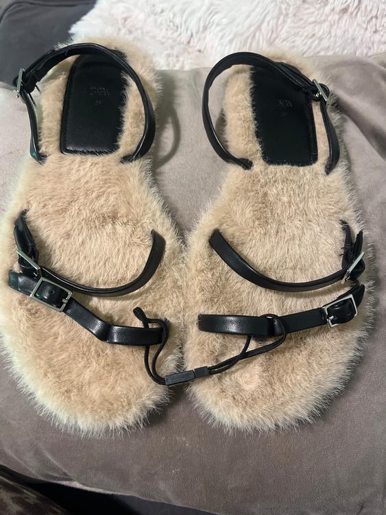 Beige plush sole sandals with black leather straps on a grey surface. White fabric textured background.