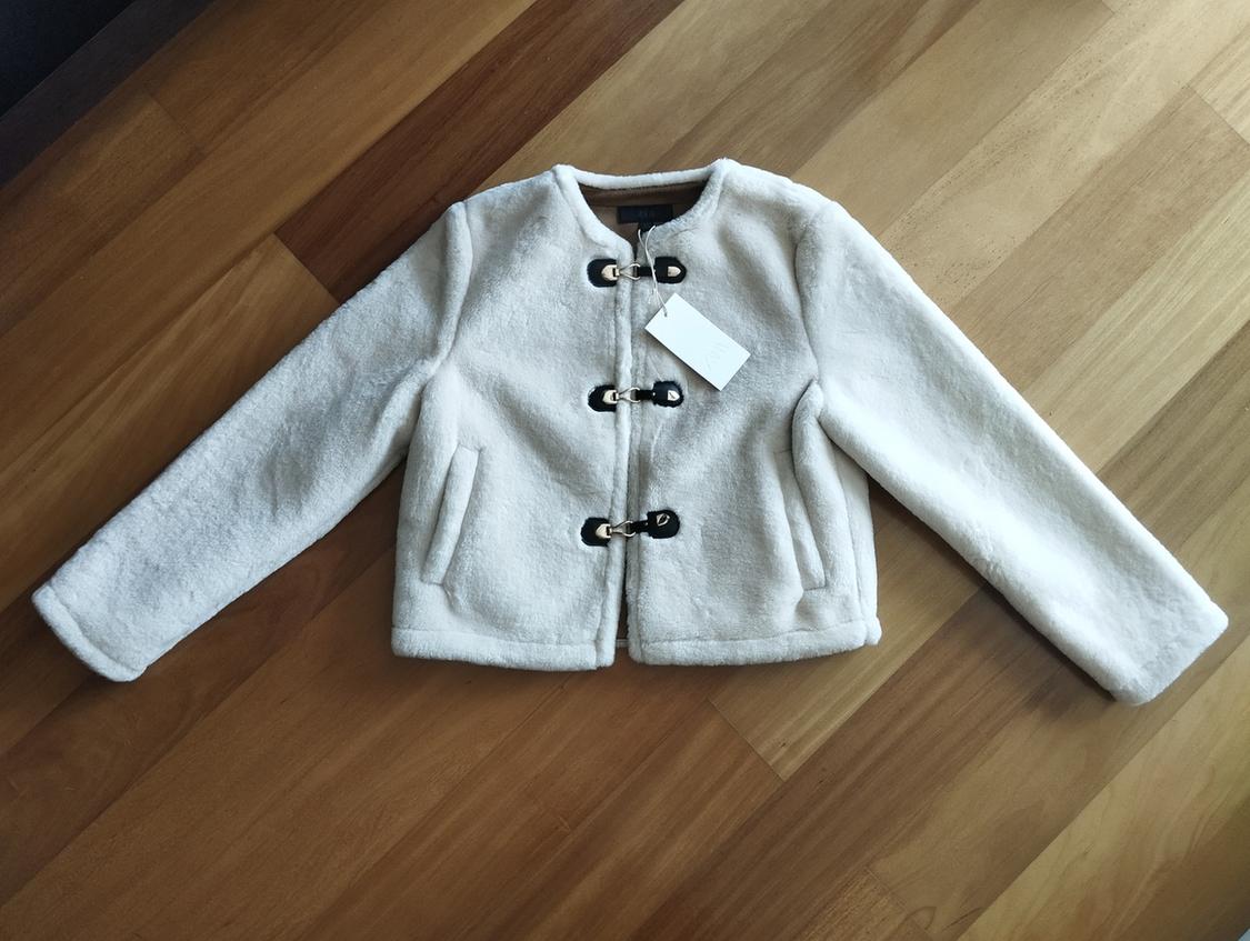 White wool jacket with black buckle fastenings on a wooden floor. White tag hanging from the collar.