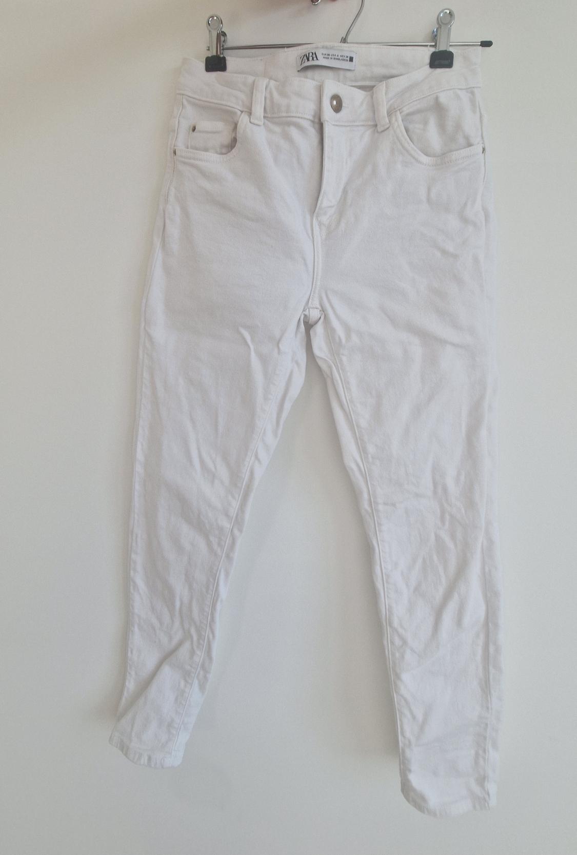 White trousers hanging on a hanger with clips, label visible at the waist, white background.