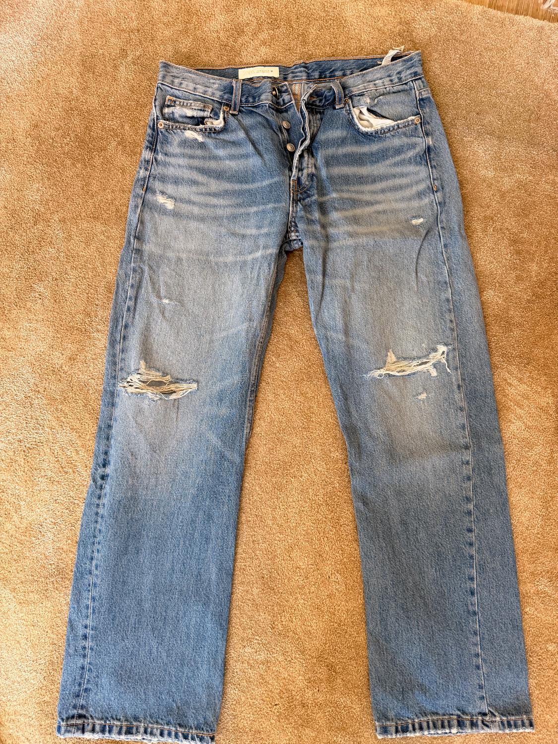 Distressed blue jeans with rips at the knees and thighs, on a beige rug.