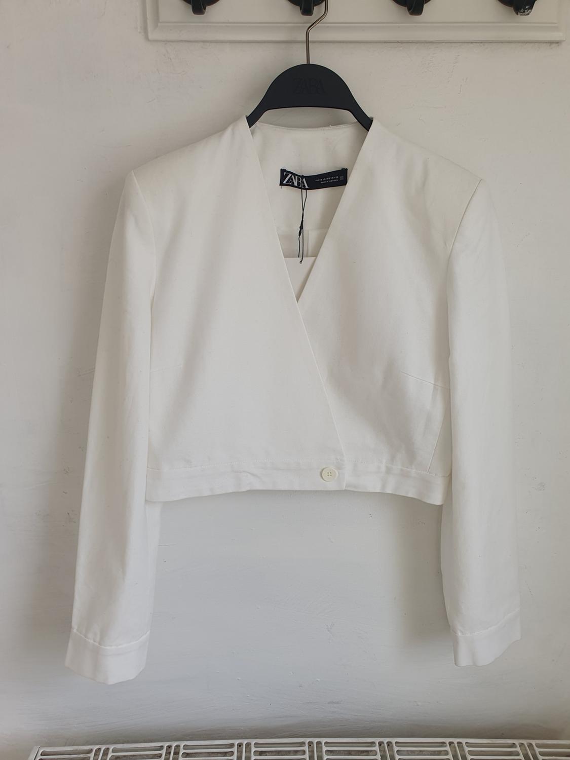 White, long-sleeved jacket hanging on a black hanger with the label visible. White background and textured wall.