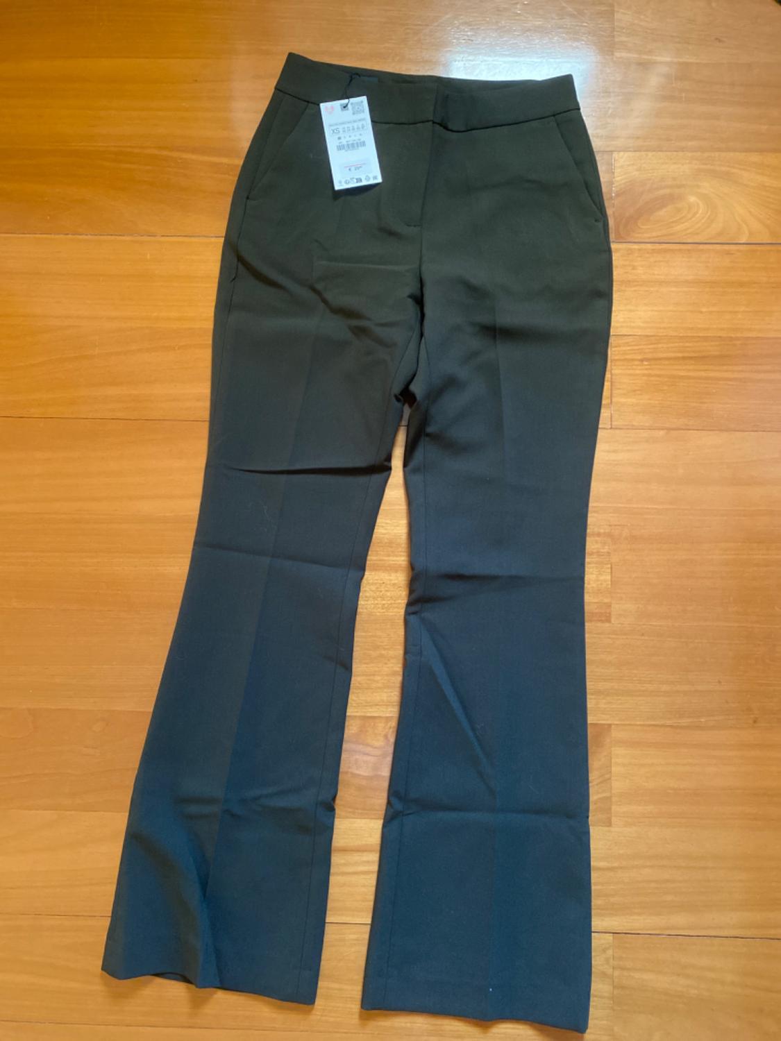 Black wide-leg trousers on a light wooden floor. White tag hanging from the right pocket.