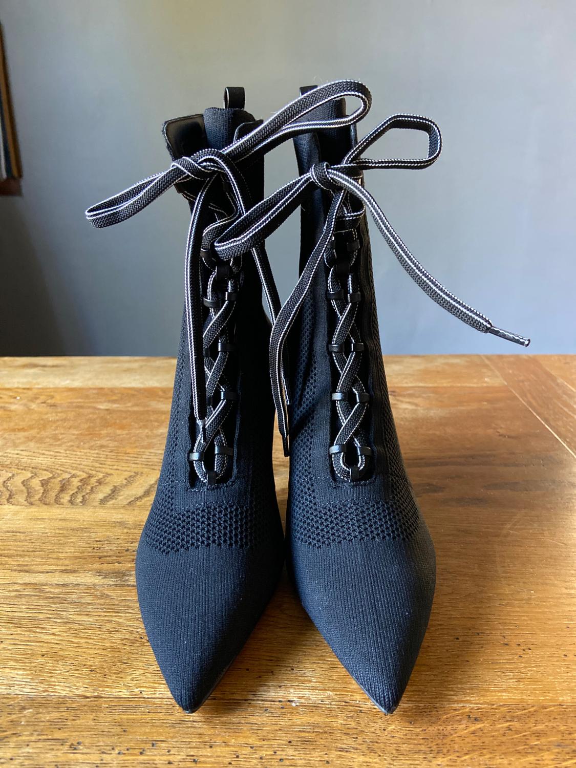 Black knitted ankle boots with a pointed toe and white lace-up
details tied in a bow, resting on a wooden table.