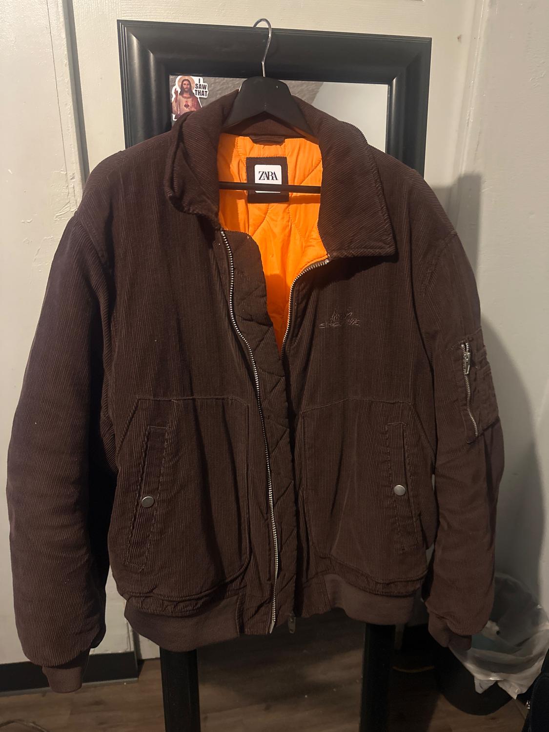 Brown corduroy jacket with an orange lining hanging on a black hanger. Visible label with the text "ZARA". Background with a black frame.
