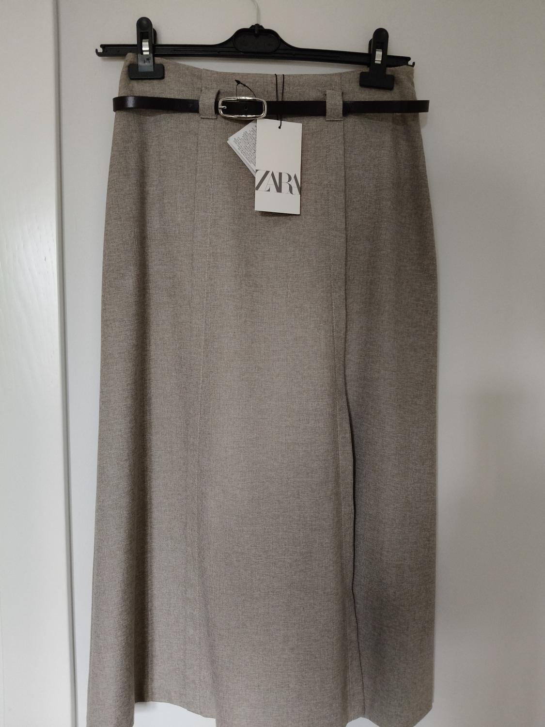 Grey fabric skirt hanging on a black hanger, with a black belt and visible Zara label. White background.
