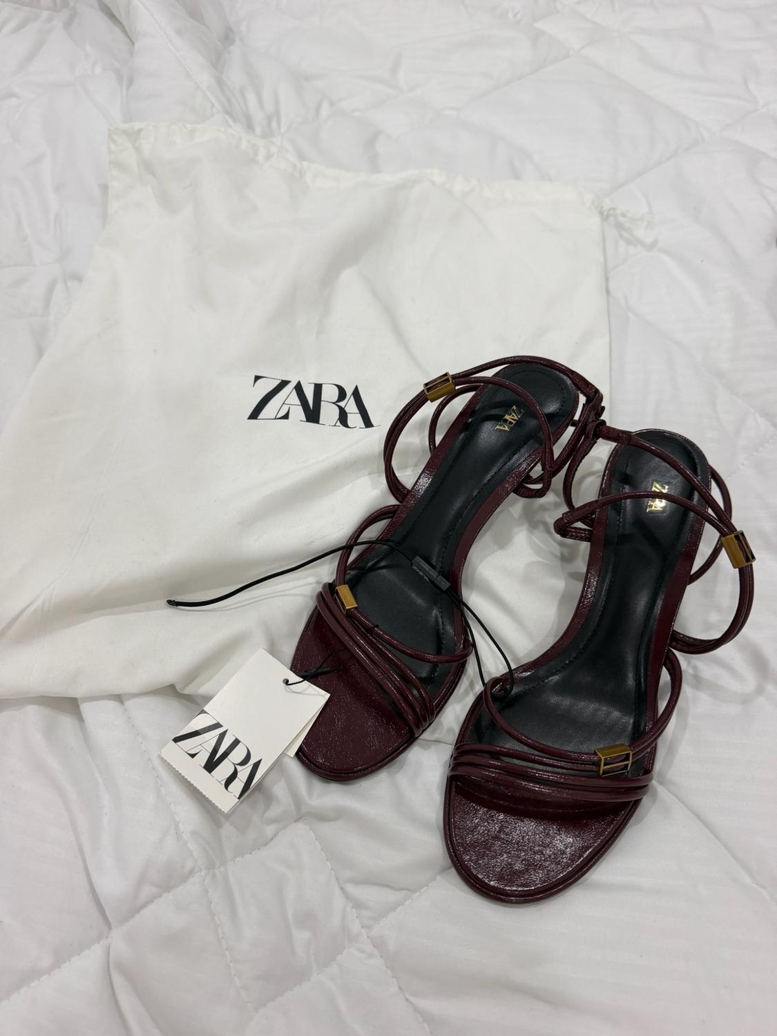 Burgundy heeled sandals with straps on padded white fabric, next to a bag and a label bearing the text ZARA.
