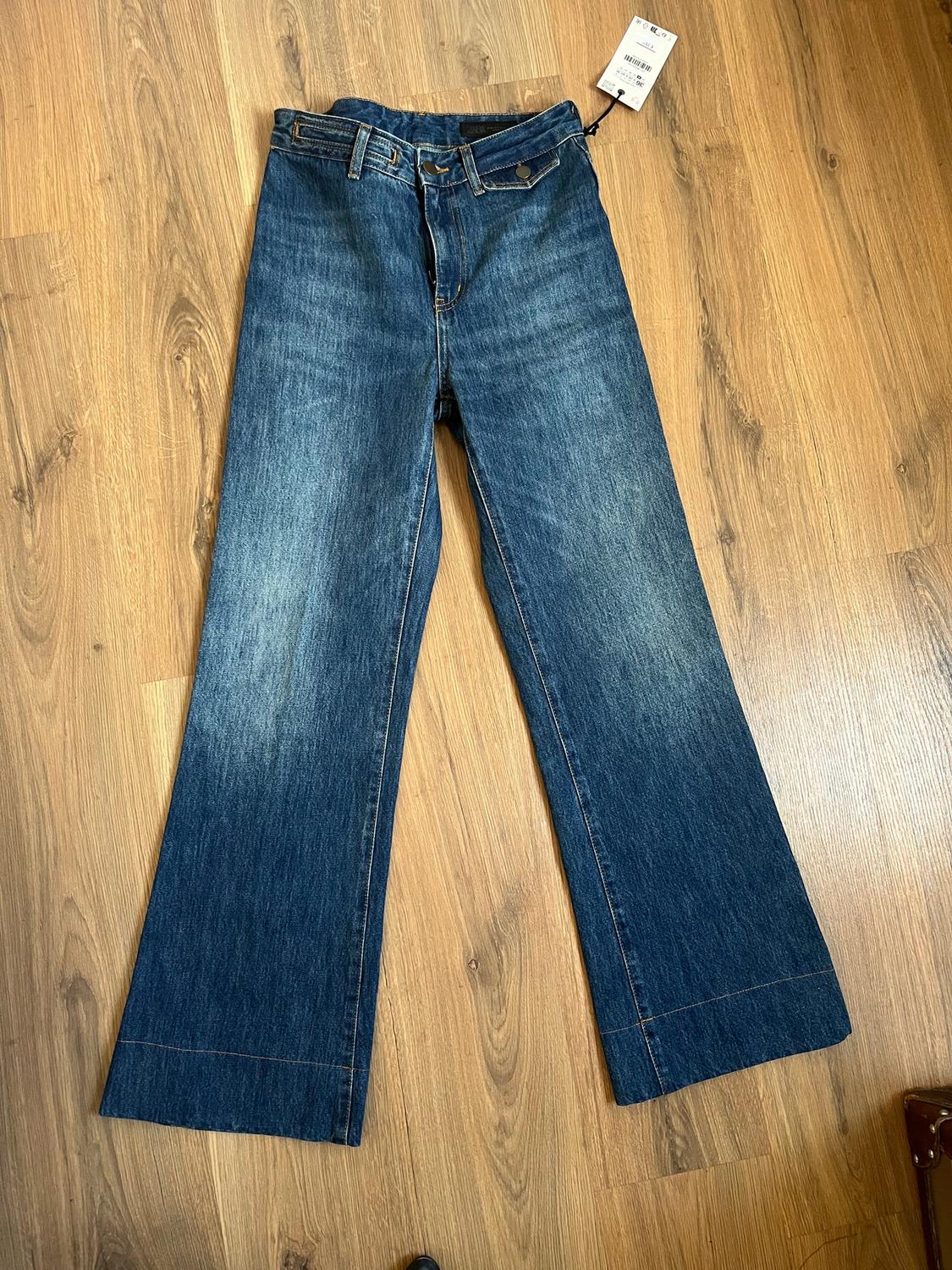 Wide-leg jeans on a light wooden floor. Visible waist label with price and size details.