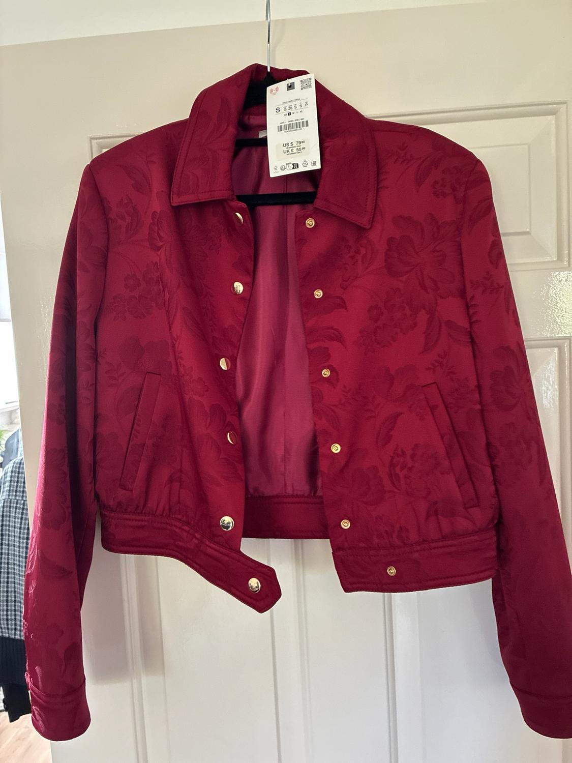 Burgundy jacket with floral texture and gold press stud buttons, hanging on a black hanger on a white door.