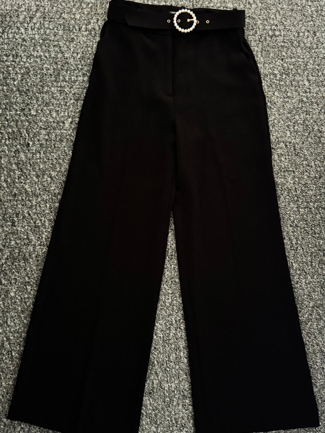 Black wide-leg trousers on a grey rug, with a black belt and buckle decorated with white pearls.