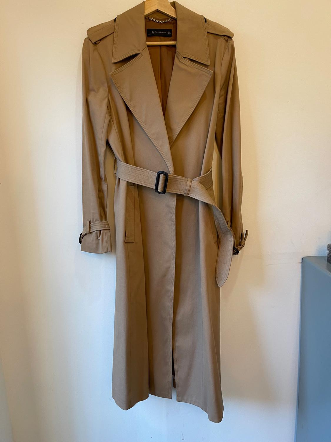 Beige trench coat hanging on a wooden hanger, with an adjustable belt and wide lapels, on a white background.