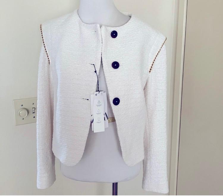 White textured fabric jacket with large black buttons on a mannequin. Tags hanging from the front. Neutral background.
