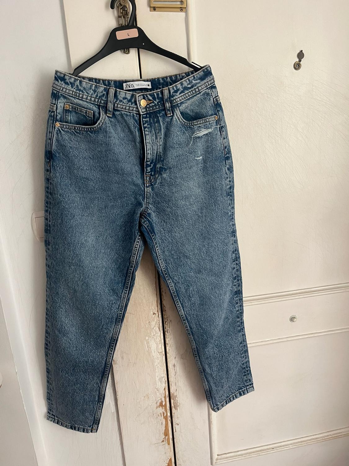 Blue denim jeans with a rip, hanging on a black hanger over a white wooden door. Visible text: ZARA and L.