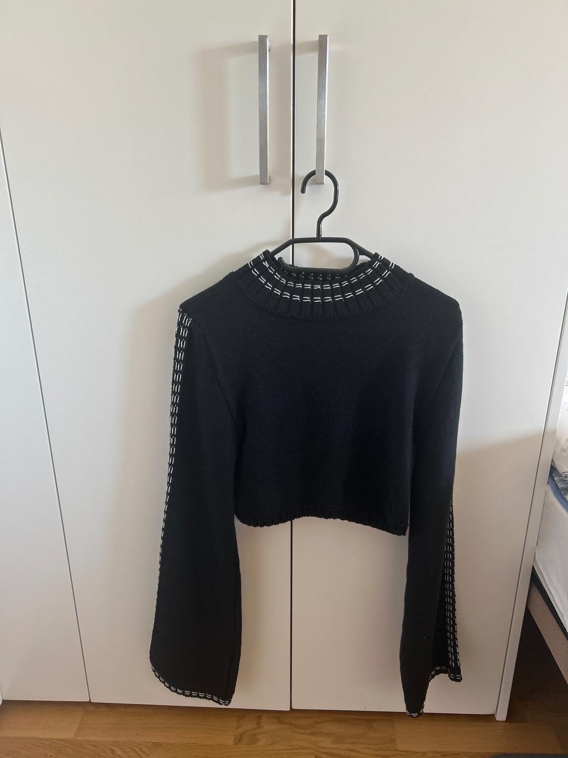 Long-sleeved black jumper with white detailing on the collar and cuffs, hanging on a hanger in front of a white wardrobe.