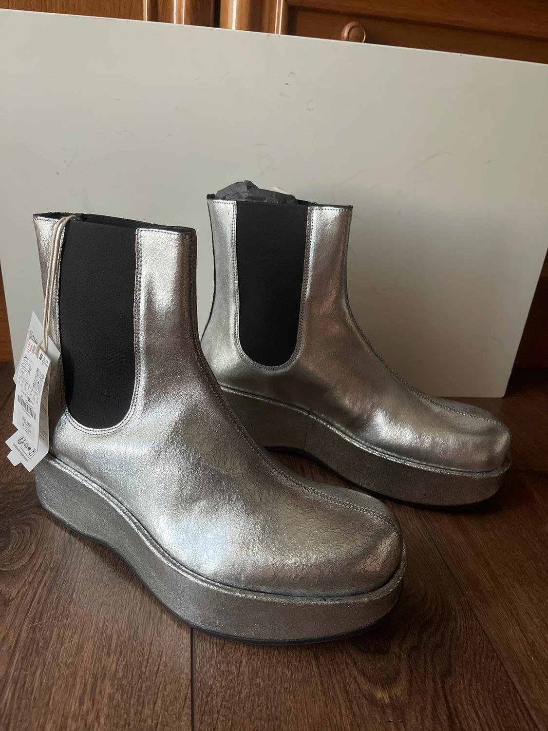 Silver platform boots with a metallic finish and black side gussets, set on a wooden floor against a white background.