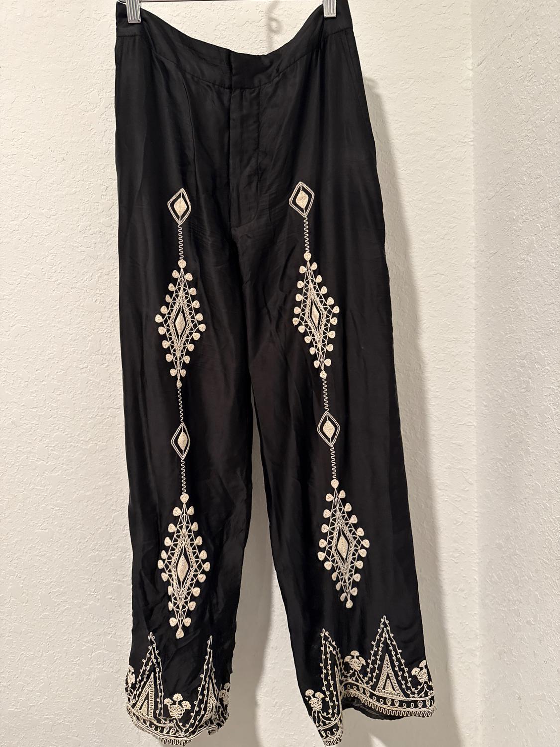 Black trousers hung with white geometric embroidery and detailing on the hems, on a white background.