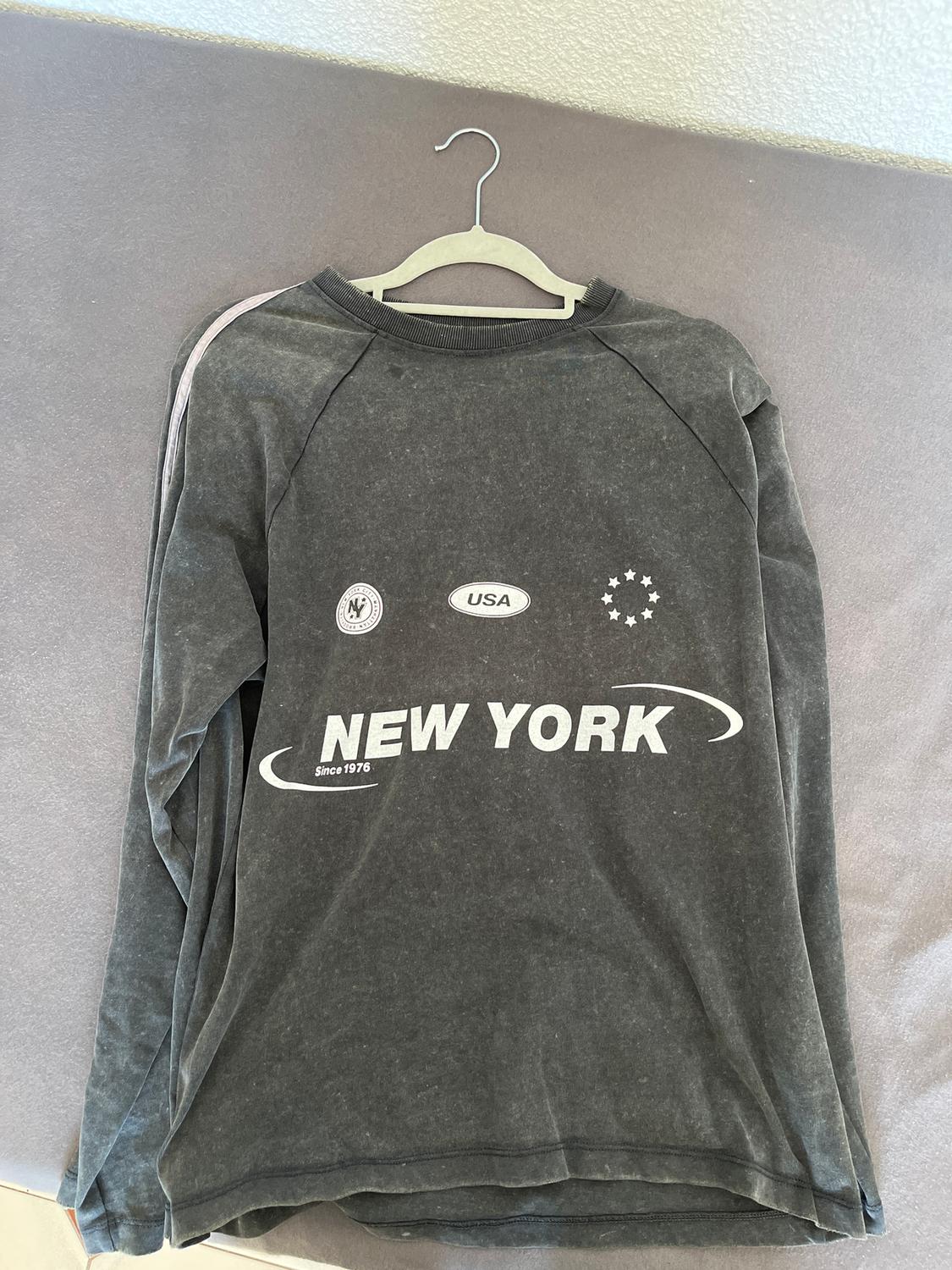 Grey sweatshirt with "NEW YORK Since 1976", "USA" text and symbols on the chest, hanging on a hanger against a grey surface.
