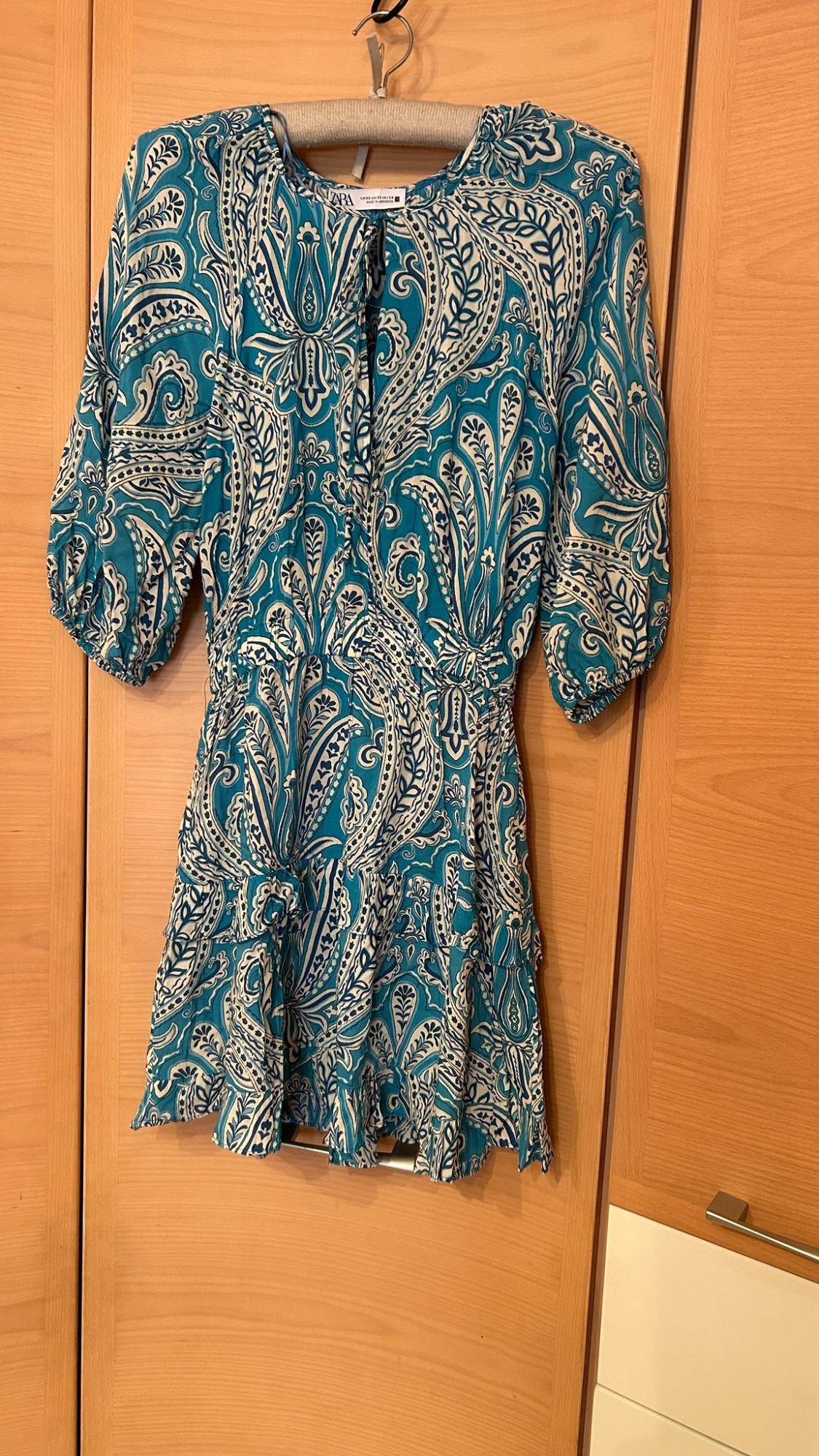 Short blue and white paisley print dress, with short sleeves and ruffles, hanging in a wooden wardrobe. Text: ZARA.