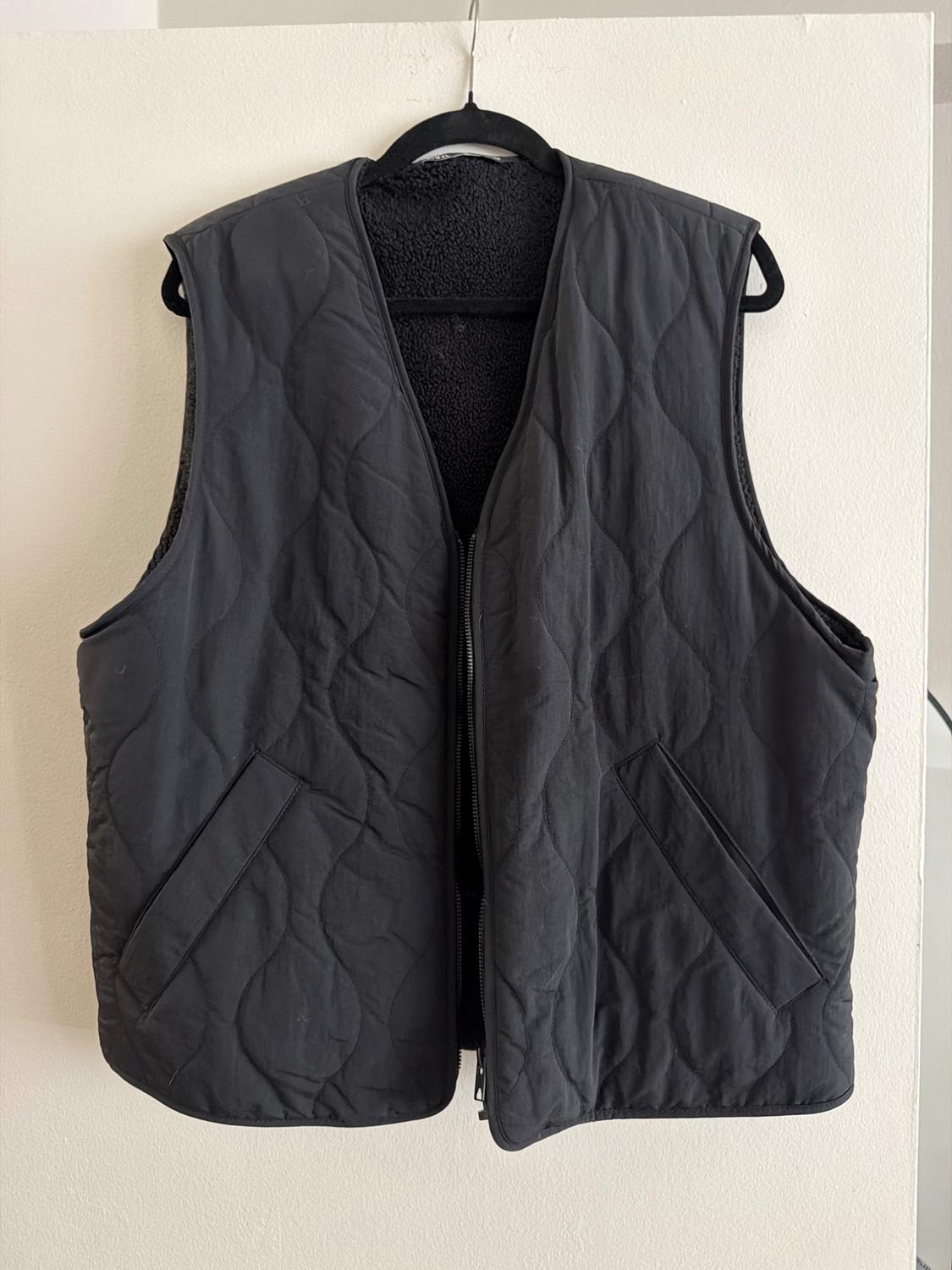 Black padded gilet hanging on a black hanger against a white background. It has slanted pockets and a zip fastening.