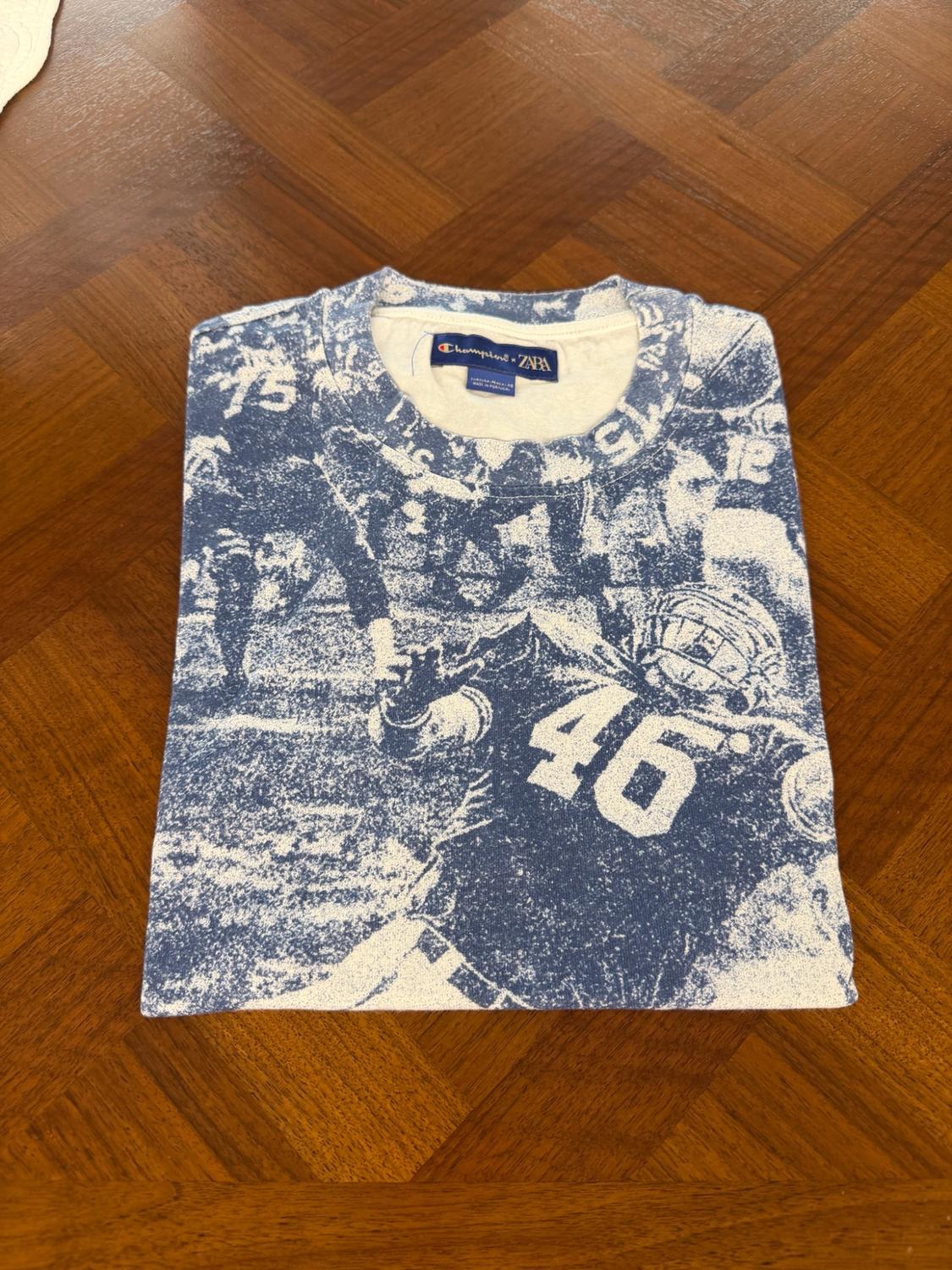 Μπλε και λευκό διπλωμένο T-shirt πάνω σε ξύλο. Σχέδιο με παίκτη αμερικανικού ποδοσφαίρου, αριθμοί 46 και 75. Ετικέτα Champion x ZARA.