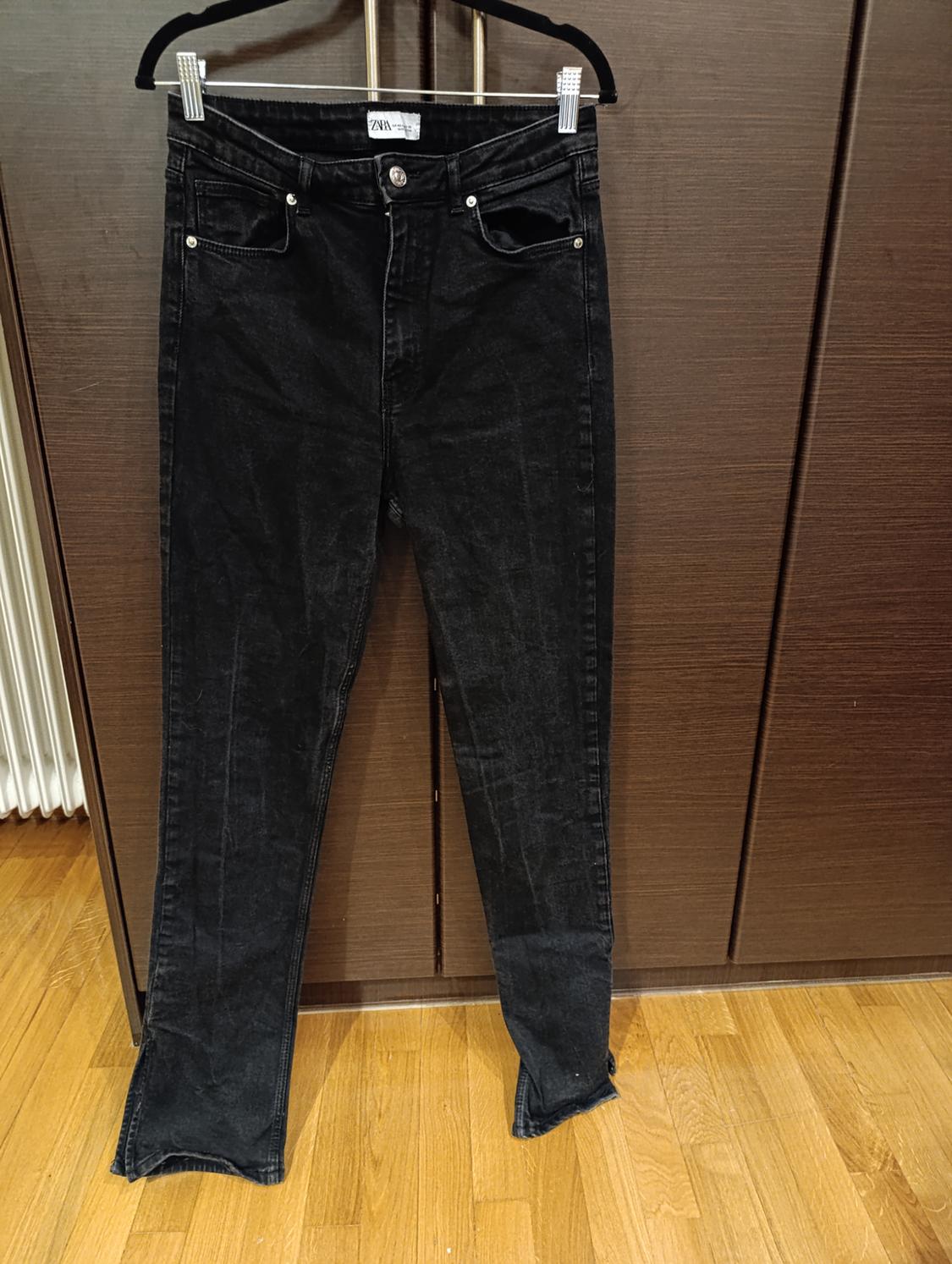 Black trousers hanging on a hanger in front of a wooden wardrobe. Label visible on the waistband. Light wooden floor.