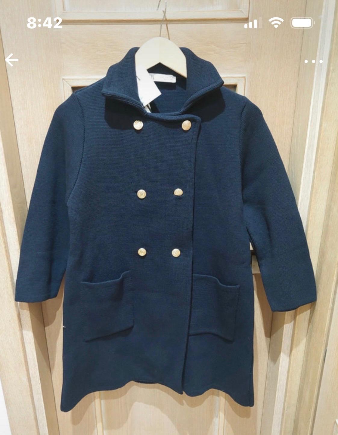 Navy blue knitted coat with double-breasted gold buttons and pockets, hanging on a wooden door. Text: 8:42.