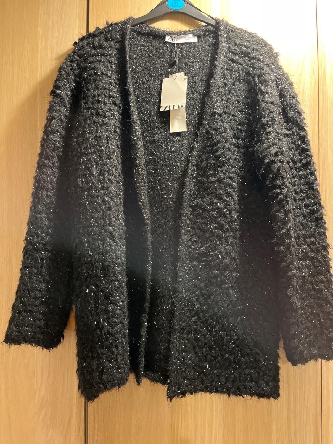 Chaqueta negra de punto grueso colgada en una percha, con etiqueta visible. Fondo de madera clara.