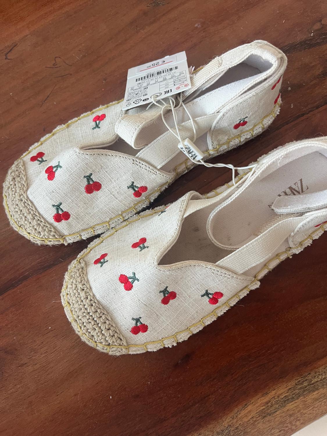 Beige canvas shoes with red cherry embroidery and a jute sole on a wooden table. Price tag visible.