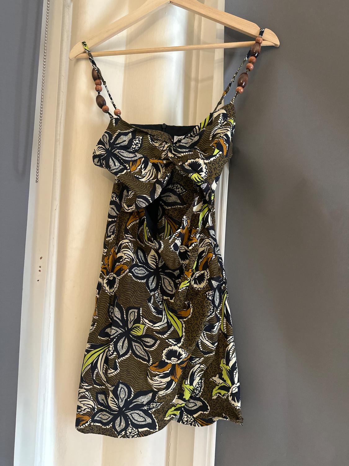 Short dress with a floral print, cut-outs and straps with wooden beads, hanging on a hanger over a white door frame.