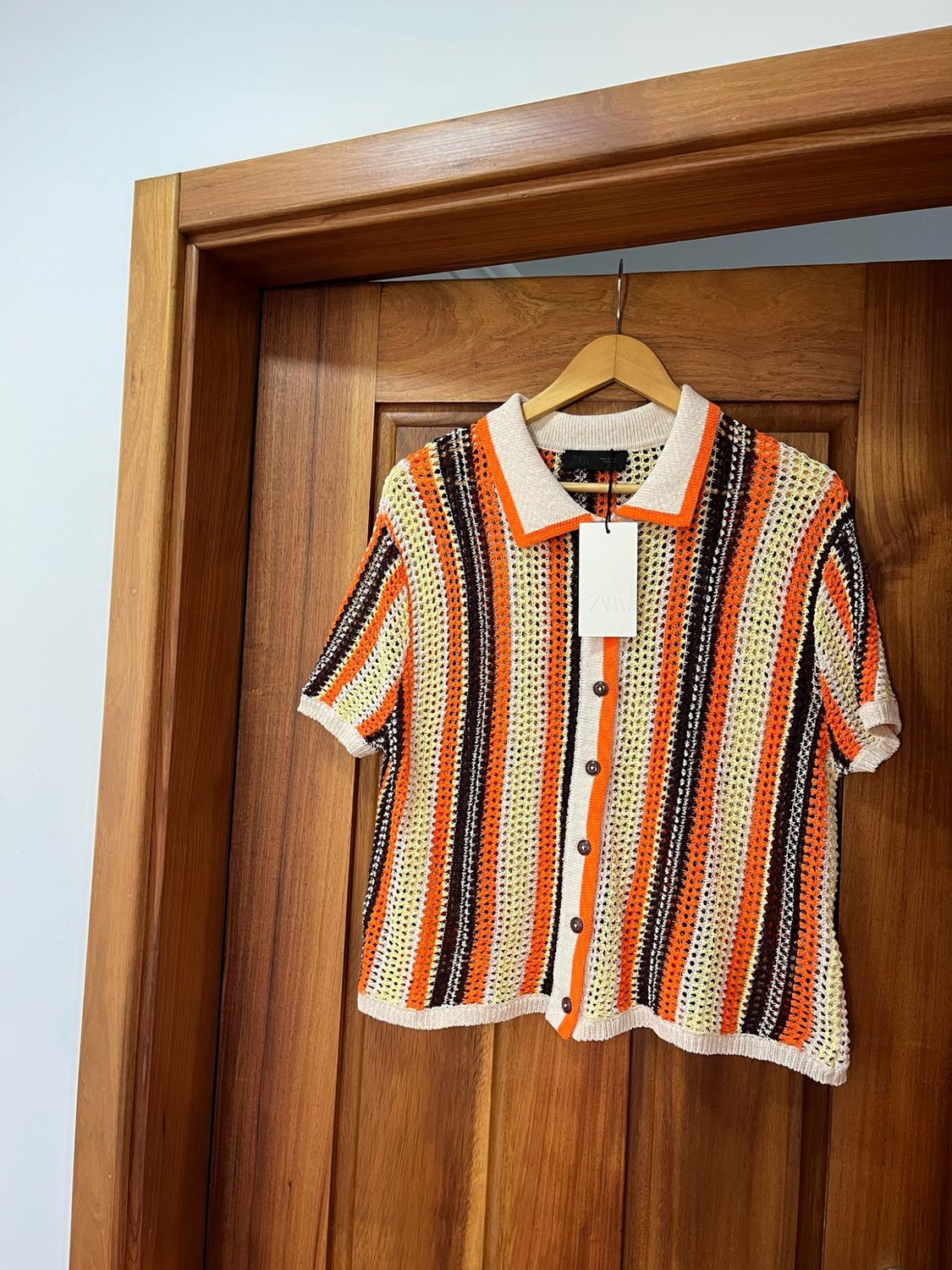 Open-knit polo shirt with orange, brown, and yellow vertical stripes, hanging on a wooden door. Label with the text ZARA.