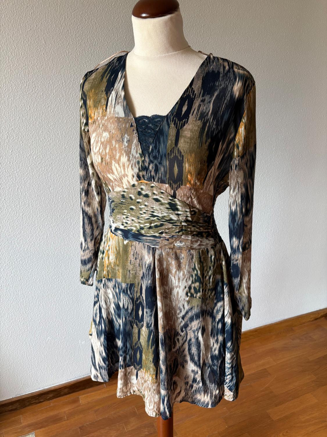 Mannequin wearing a long-sleeved dress with an abstract print in earthy tones, a fitted waist and a V-neck with lace detailing. White wall and wooden floor.
