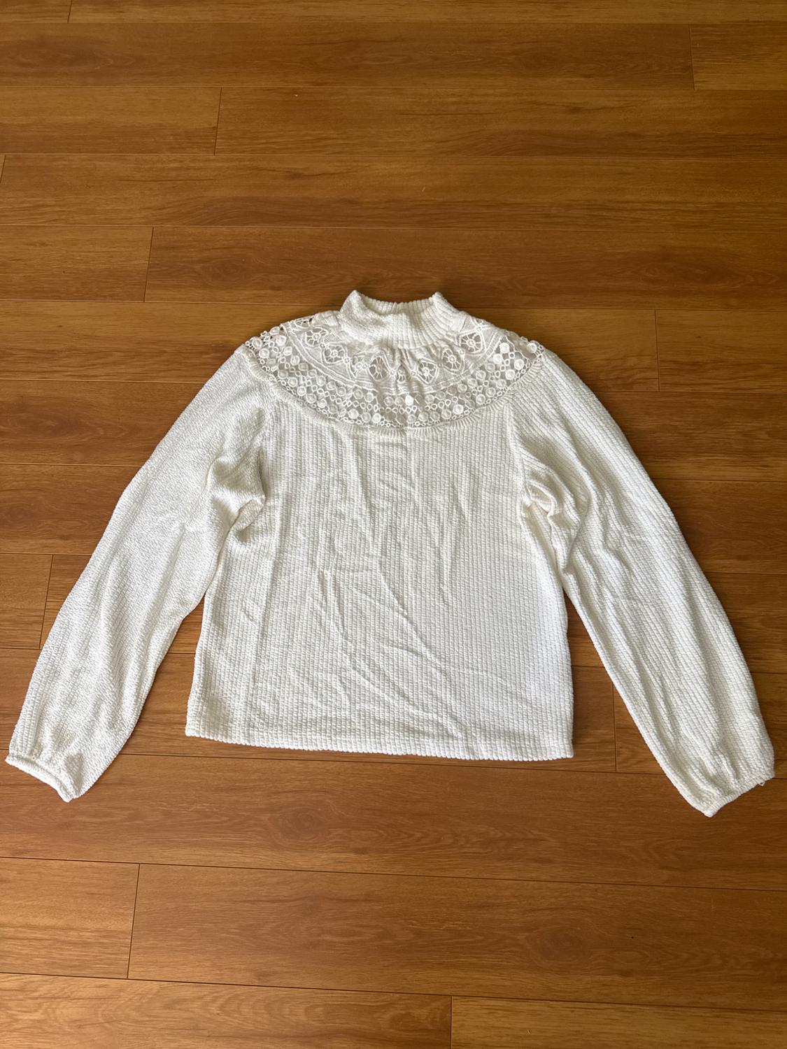 White long-sleeved polo neck jumper with lace detail on the shoulders, on a brown wooden floor.