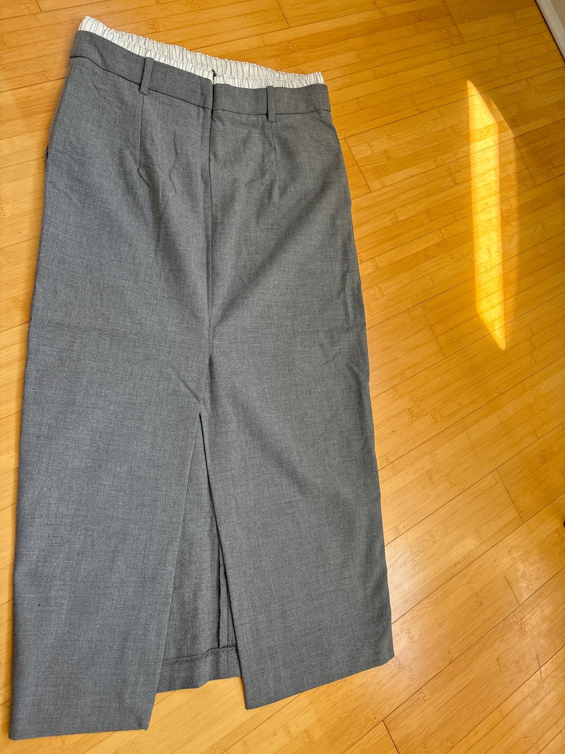 Light gray fabric skirt with a front slit, visible white elastic waistband, on a light wood floor illuminated by natural light.