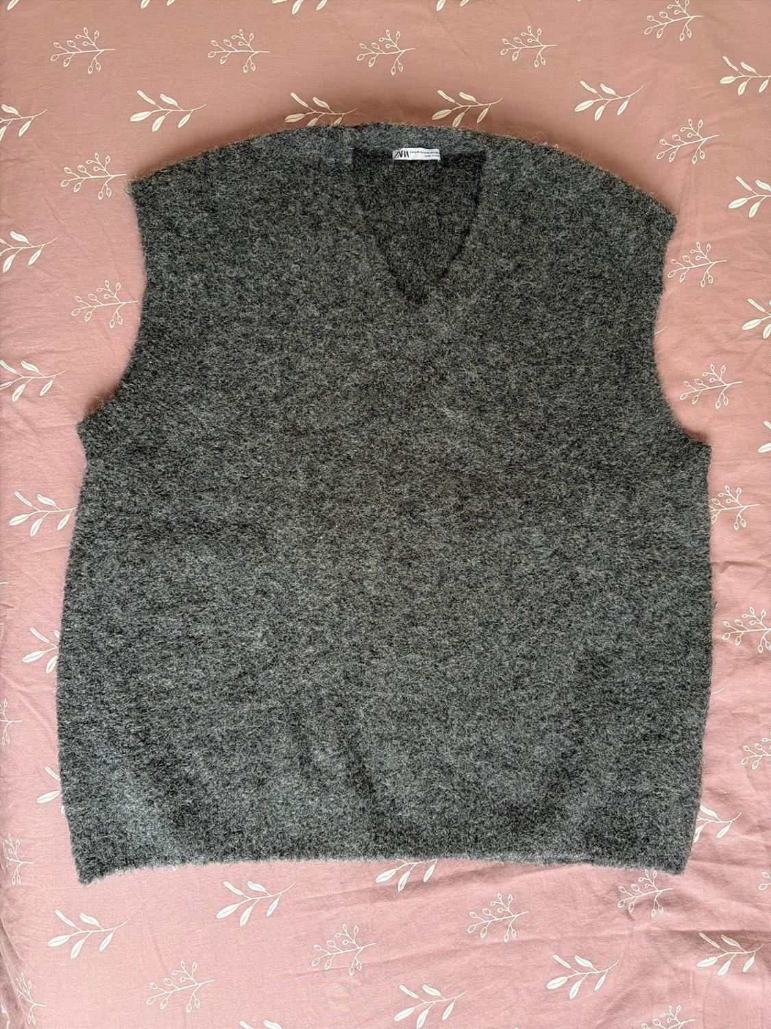 Dark gray knit V-neck vest over a pink fabric background with a white leaf print. ZARA label visible on the collar.
